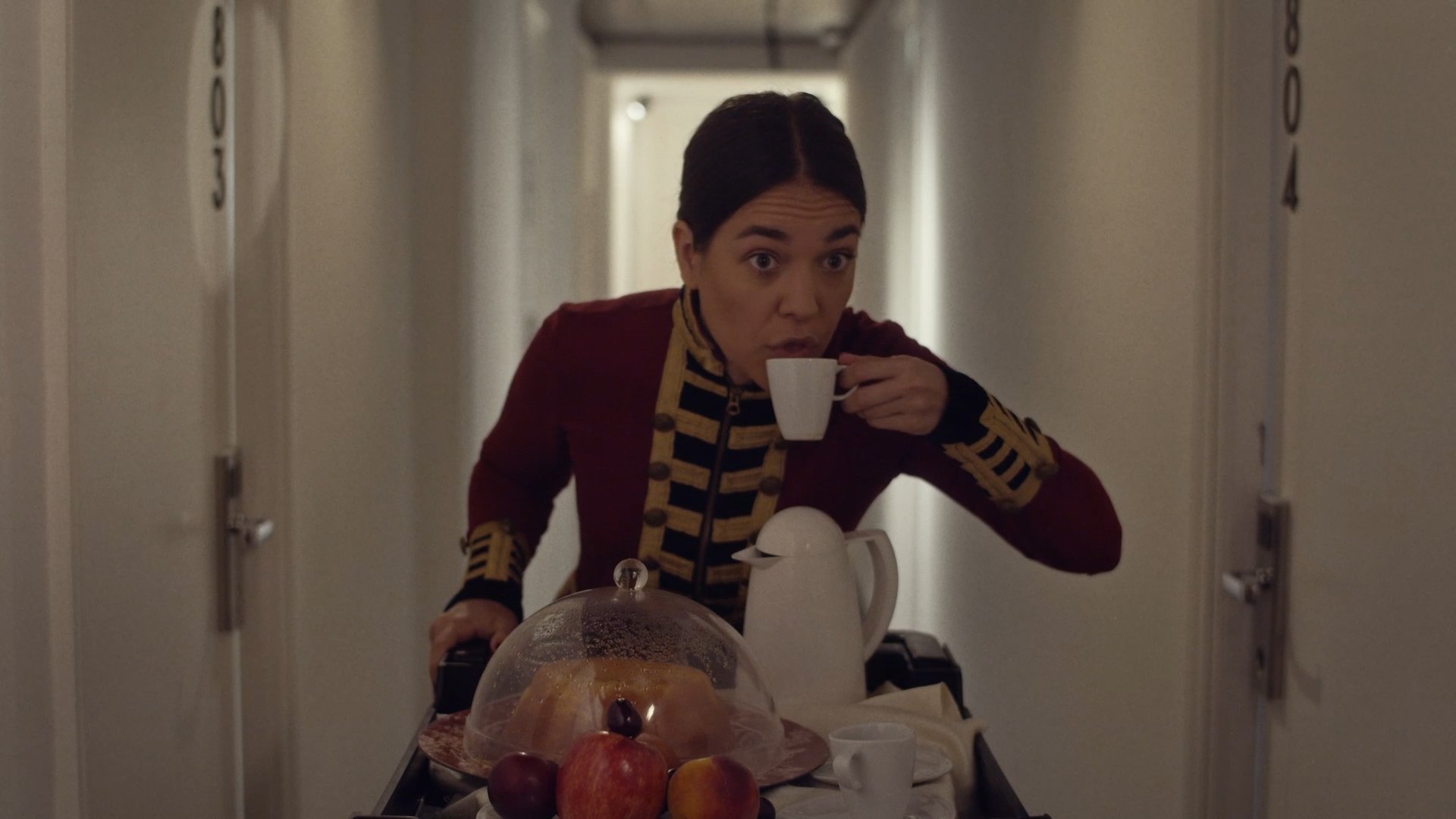 Bellhop in a red braided jacket sipping coffee behind a room-service cart